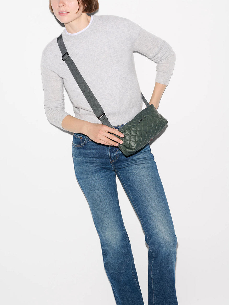 A person wearing a light grey sweater, blue jeans, and the MZ Wallace Metro East West Crossbody in Juniper stands against a plain white background.