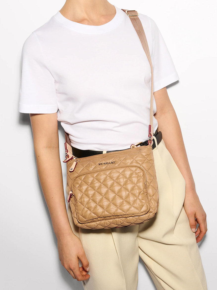 A person in a white T-shirt and beige pants is carrying the MZ Wallace Metro Scout Deluxe crossbody bag in Caramel Oxford, featuring an adjustable strap and branded logo.