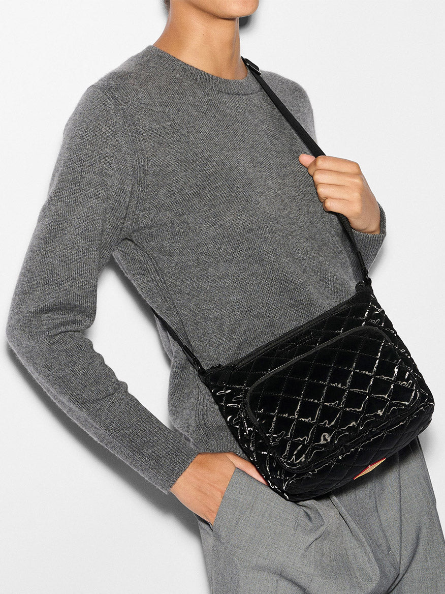 A person in a gray sweater and pants confidently holds a black quilted shoulder bag, showcasing the elegant design of the MZ Wallace Metro Scout Deluxe in Black Liquid Oxford.