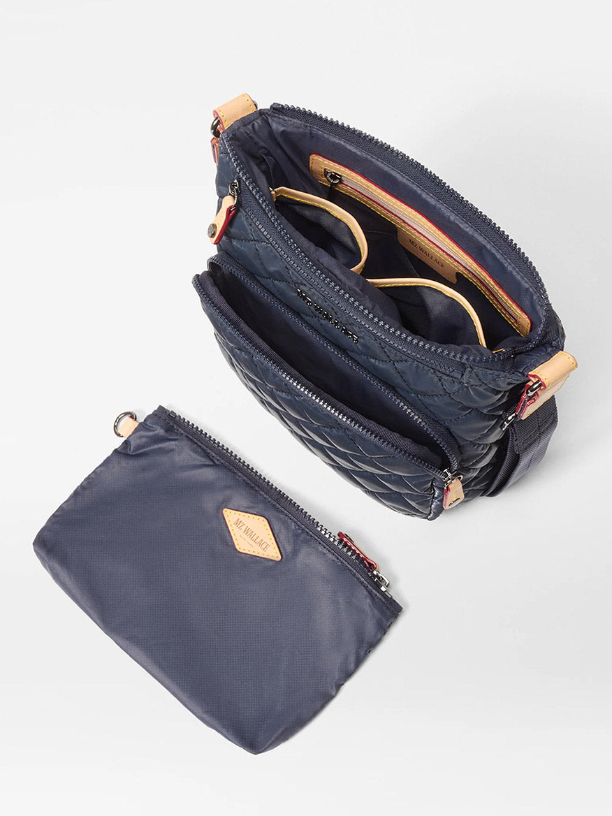 The MZ Wallace Metro Scout Deluxe in Ink Liquid Oxford, shown open with glasses inside and next to a matching detachable zip pouch, features multiple compartments and a stylish crossbody design.