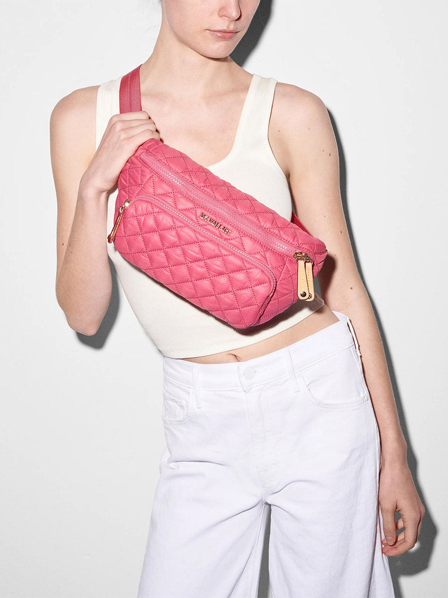 Woman wearing a white tank top and white pants, holding a pink quilted MZ Wallace Metro Sling in Zinnia Oxford with an adjustable strap and gold zippers.