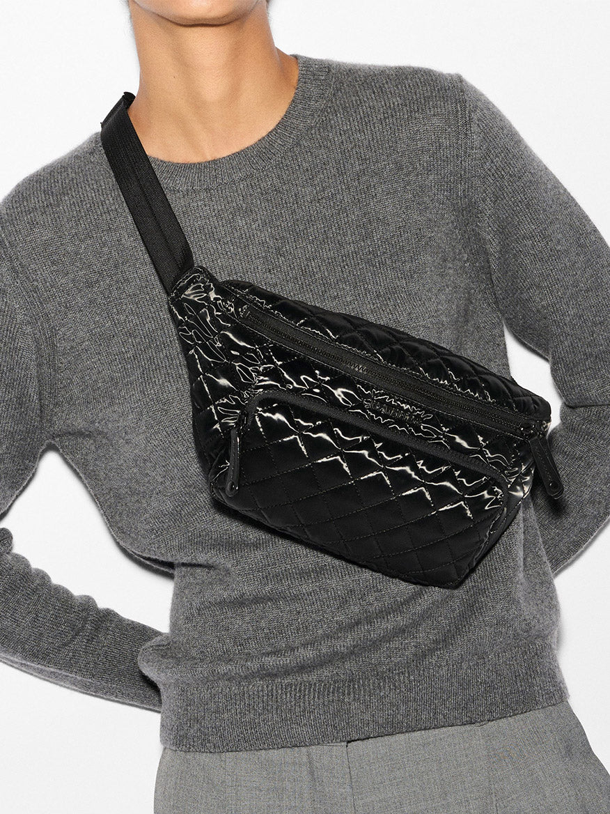 Wearing a gray sweater and pants, a person finishes their outfit with a black Metro Sling in Black Liquid Oxford by MZ Wallace, which offers an adjustable strap for hands-free convenience.