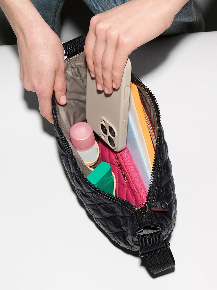 A person puts a smartphone into the MZ Wallace Metro Smile Bag in Black Oxford, trimmed with Italian leather, alongside a lipstick, lotion bottle, and colorful zippered pouches.