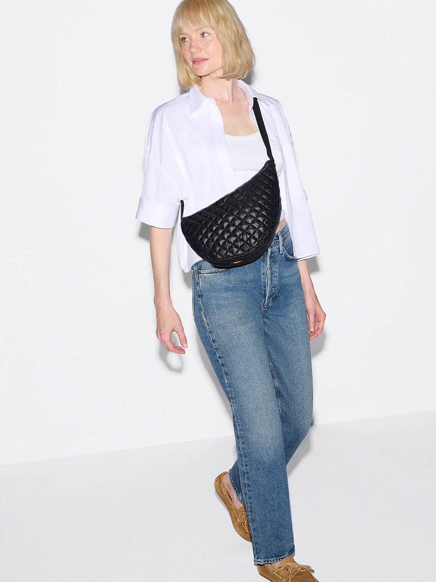 A woman with blonde hair wears a white shirt, blue jeans, tan shoes, and carries the MZ Wallace Metro Smile Bag in Black Oxford, featuring Italian leather trim and an adjustable nylon strap, standing against a white background.