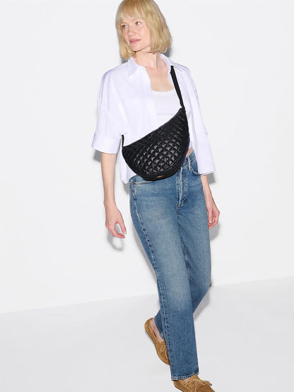 A woman with blonde hair wears a white shirt, blue jeans, tan shoes, and carries the MZ Wallace Metro Smile Bag in Black Oxford, featuring Italian leather trim and an adjustable nylon strap, standing against a white background.
