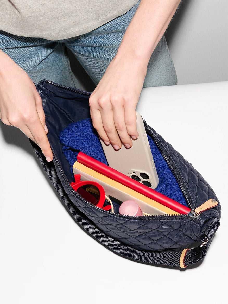 A person places a smartphone into the MZ Wallace Metro Smile Bag in Dawn Oxford, which contains a blue cloth, red sunglasses, a makeup compact, and a yellow and red item.