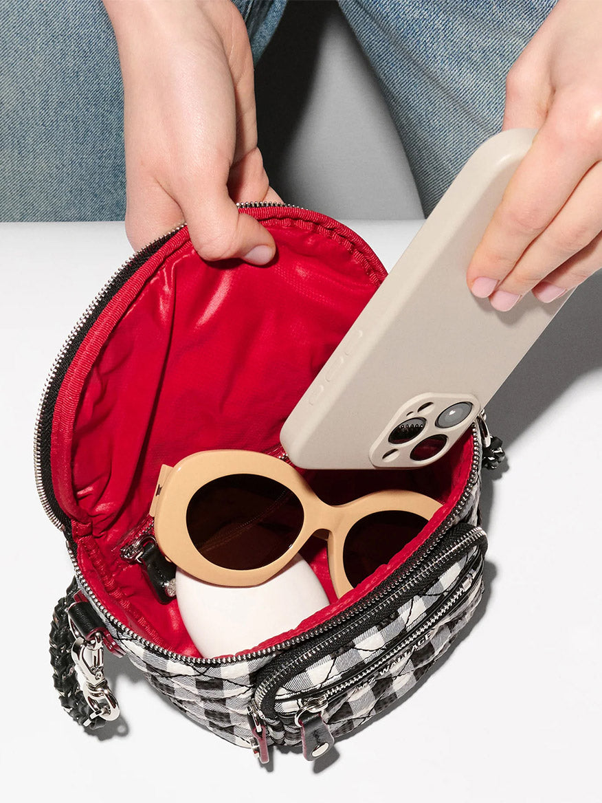 A person places a smartphone into an MZ Wallace Micro Crosby Crossbody Sling in Black/White Gingham Oxford, which also holds beige sunglasses and a white object.