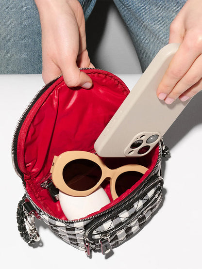 A person places a smartphone into an MZ Wallace Micro Crosby Crossbody Sling in Black/White Gingham Oxford, which also holds beige sunglasses and a white object.