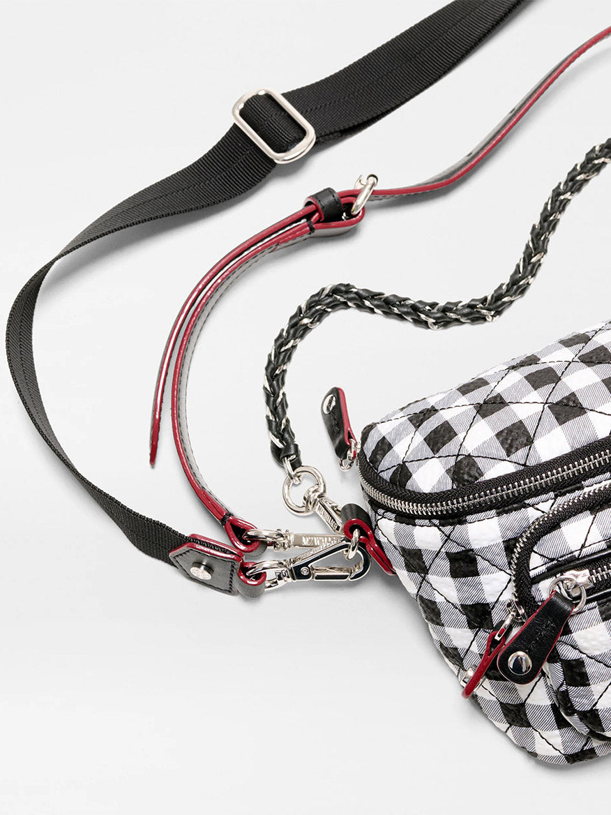 A close-up of the MZ Wallace Micro Crosby Crossbody Sling in Black/White Gingham Oxford, featuring multiple zippers, metal clasps, and adjustable black and red straps on a light background.