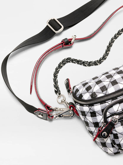 A close-up of the MZ Wallace Micro Crosby Crossbody Sling in Black/White Gingham Oxford, featuring multiple zippers, metal clasps, and adjustable black and red straps on a light background.
