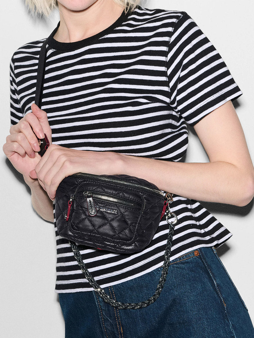 A person wearing a black and white striped shirt, holding an MZ Wallace Micro Crosby Crossbody Sling in Black Oxford, featuring Italian leather trim, silver zippers, and a chain strap.