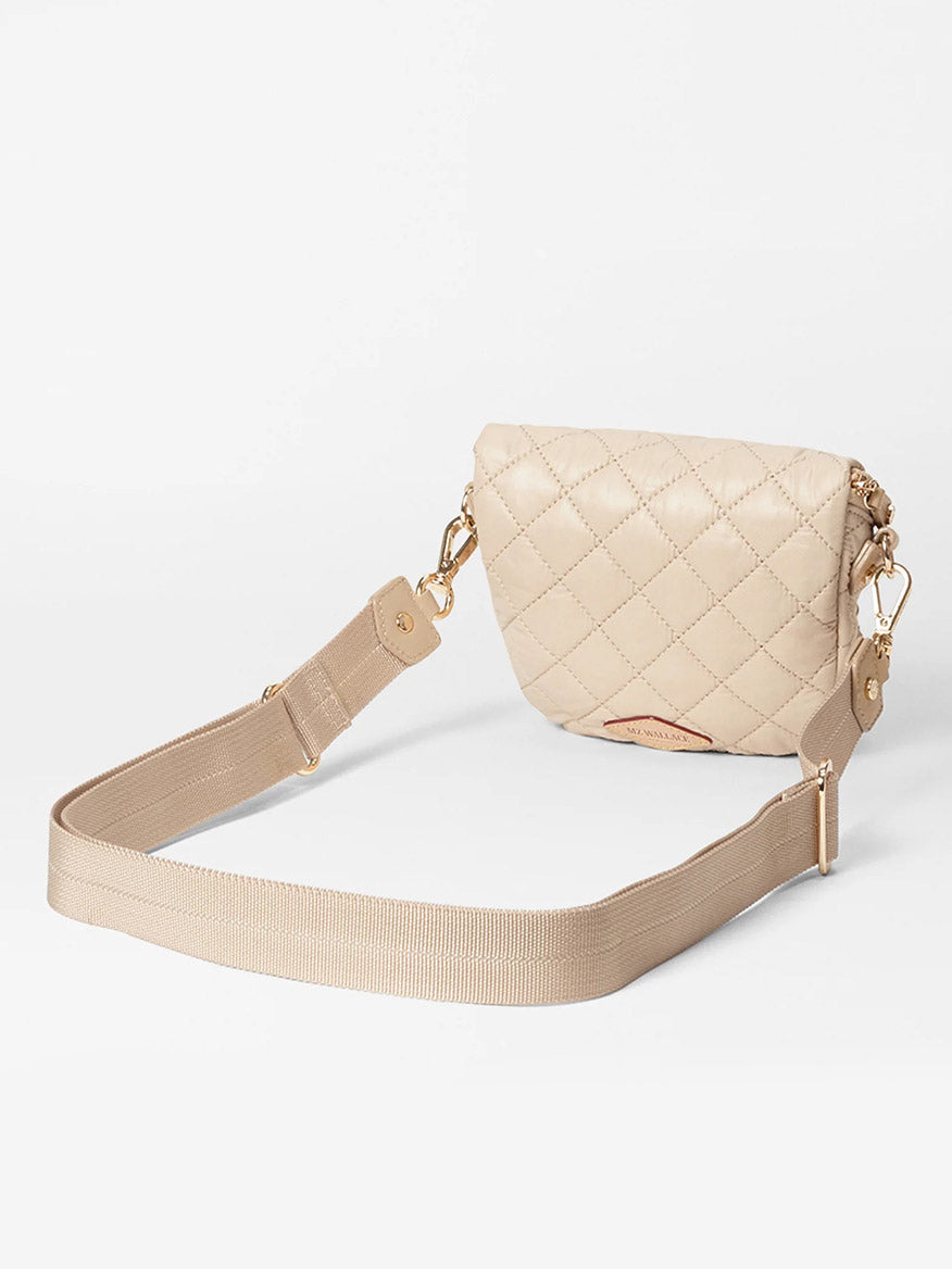 The MZ Wallace Micro Crosby Crossbody Sling in Buff Oxford, a small beige quilted bag with gold hardware and an adjustable nylon crossbody strap, rests gracefully on a plain background.