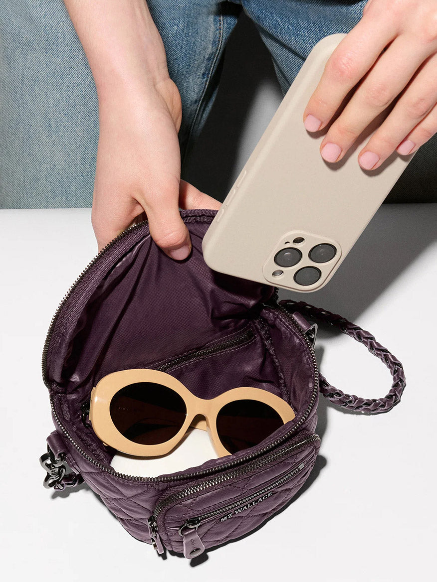 A person places a beige smartphone into a small purple recycled nylon bag, which also contains a pair of beige-framed sunglasses. The MZ Wallace Micro Crosby Crossbody Sling in Raisin Oxford is perfect for compact hands-free carry.