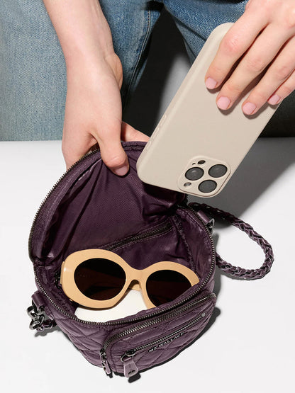 A person places a beige smartphone into a small purple recycled nylon bag, which also contains a pair of beige-framed sunglasses. The MZ Wallace Micro Crosby Crossbody Sling in Raisin Oxford is perfect for compact hands-free carry.