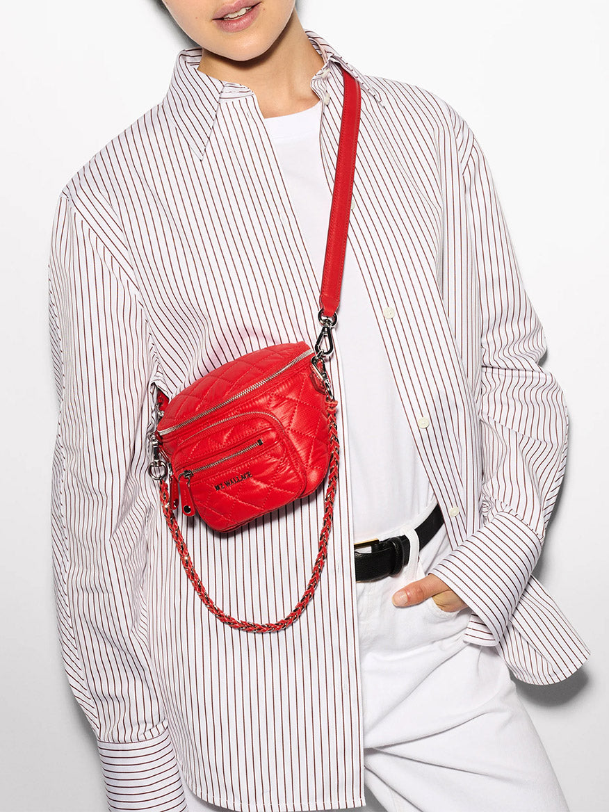 A person wears a white striped shirt with white pants and a red MZ Wallace Micro Crosby Crossbody Sling in Bright Cherry Bedford. The shirt is buttoned up with loose cuffs, complementing the stylish detachable crossbody strap of the bag.