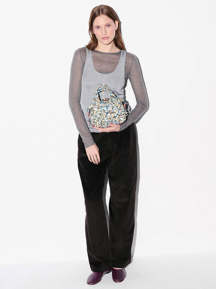 A woman stands against a white background wearing a sheer gray top, loose black pants, purple shoes, and holding the MZ Wallace Micro Sutton in Club Paint Oxford crossbody bag crafted from Liberty Fabric.