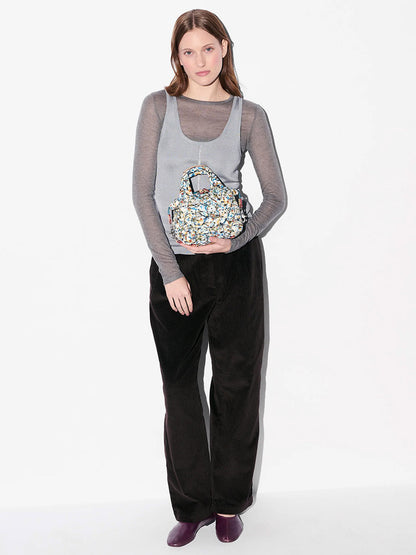 A woman stands against a white background wearing a sheer gray top, loose black pants, purple shoes, and holding the MZ Wallace Micro Sutton in Club Paint Oxford crossbody bag crafted from Liberty Fabric.