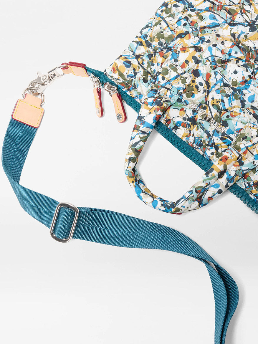 A close-up of the MZ Wallace Micro Sutton in Club Paint Oxford, a patterned foldable crossbody bag with a blue adjustable shoulder strap and dual zipper pulls, shown against a white background.
