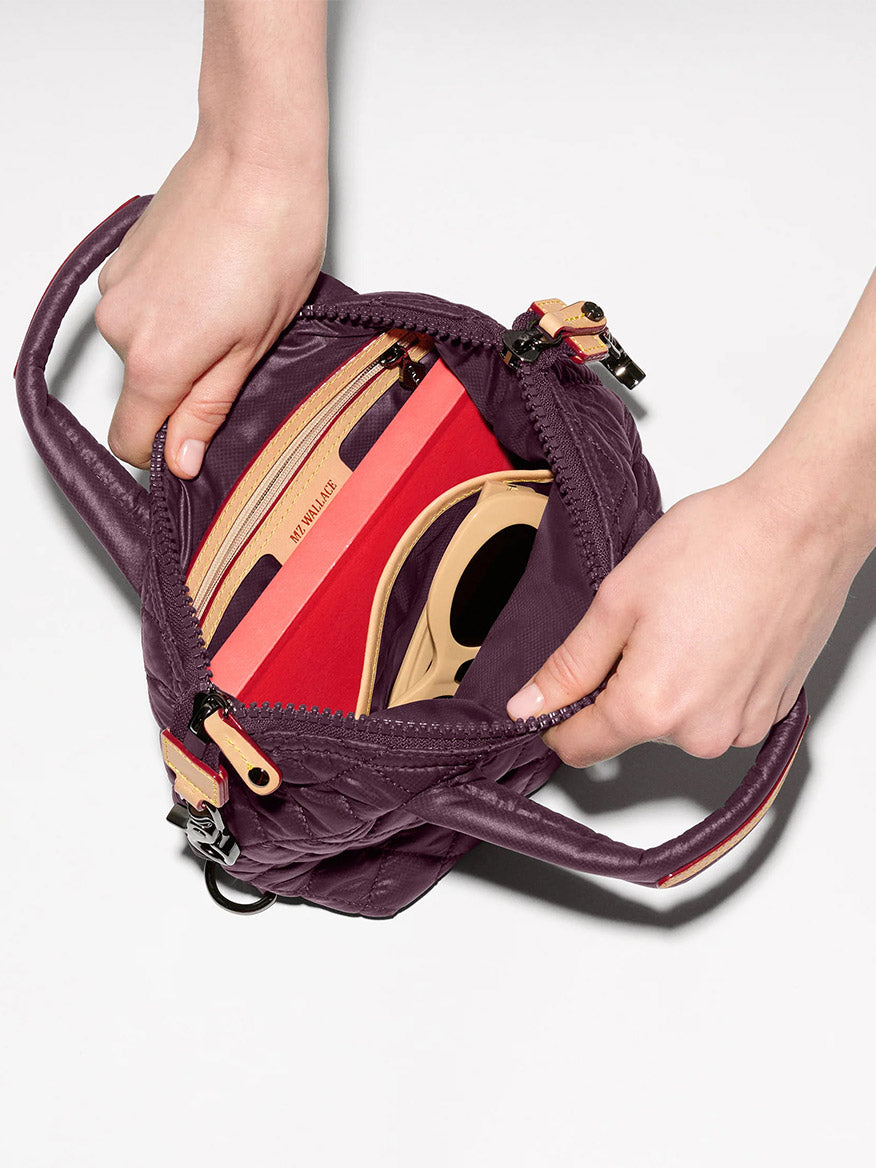 Someone opens an MZ Wallace Micro Sutton in Raisin Oxford, a compact crossbody bag made from recycled nylon, revealing beige and red interior compartments—one holding a smartphone.