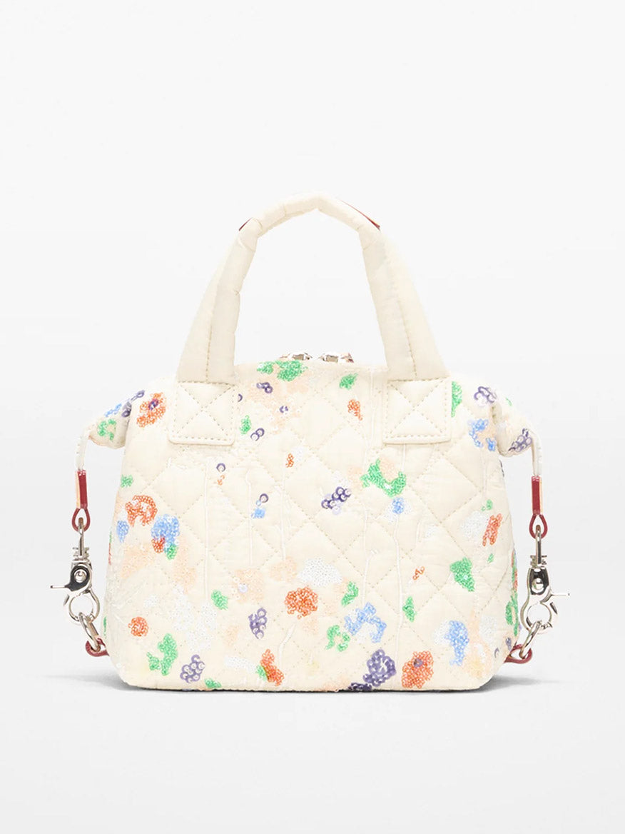 The MZ Wallace Micro Sutton in Eden Sequin Oxford is a small, quilted cream handbag featuring colorful floral patterns, two short handles, and a detachable red-and-silver crossbody strap.