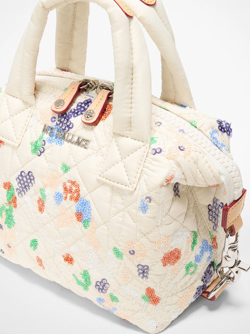 The MZ Wallace Micro Sutton in Eden Sequin Oxford is a cream quilted travel handbag featuring dual handles, multicolored floral embroidery, a zipper closure, and a metal logo plate.