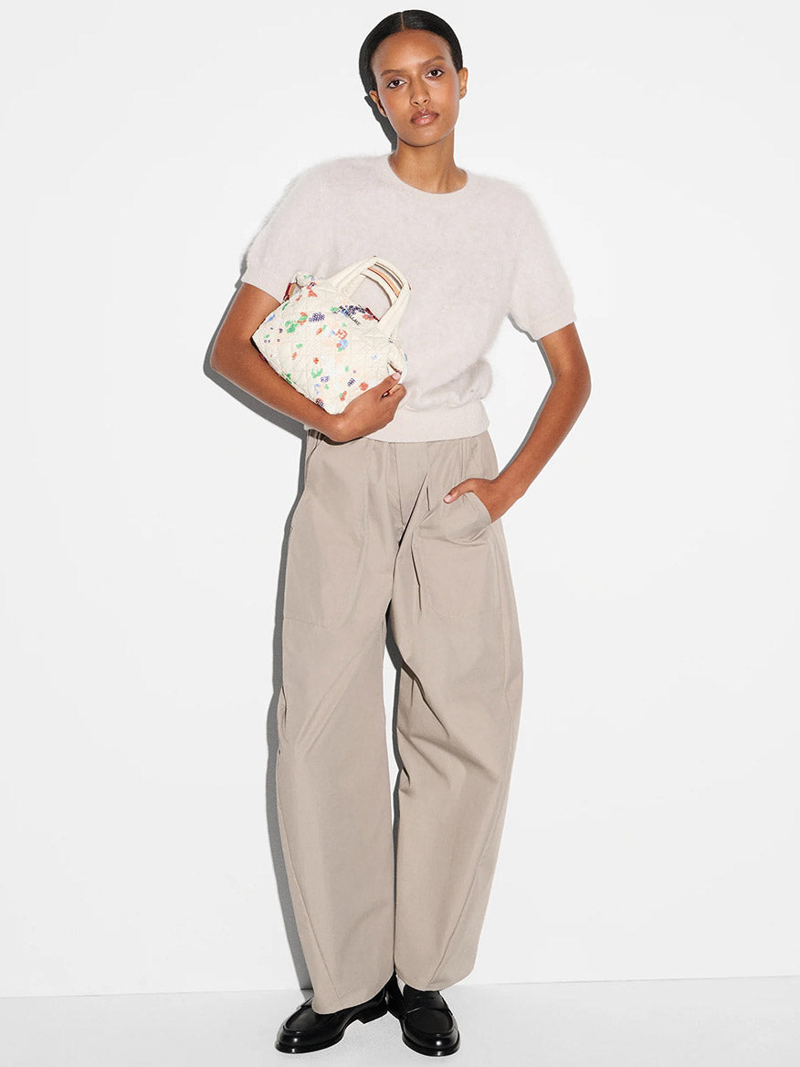 A woman stands against a white background wearing a white short-sleeve sweater, beige wide-leg pants, black shoes, and holding the MZ Wallace Micro Sutton in Eden Sequin Oxford handbag.