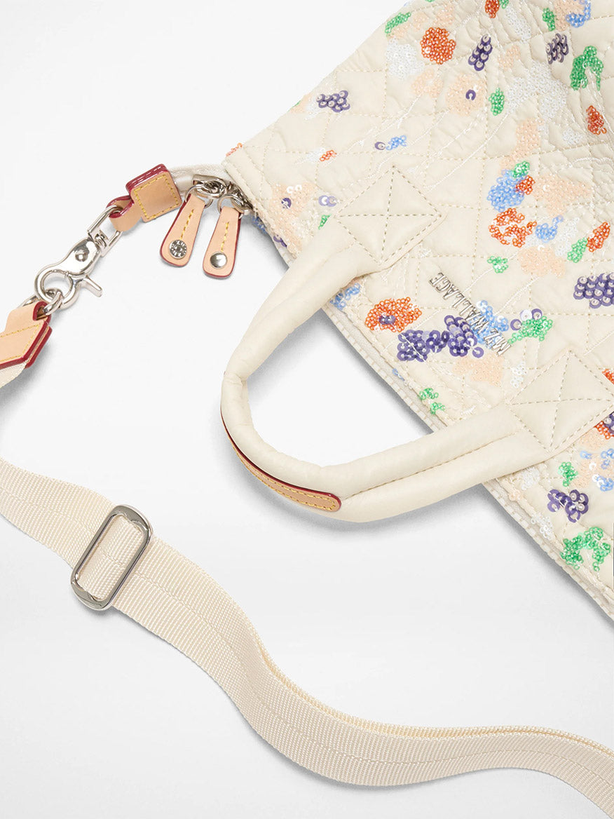 The MZ Wallace Micro Sutton in Eden Sequin Oxford is a cream quilted foldable crossbody bag with colorful floral embroidery, two handles, a detachable cream strap with silver hardware, shown on a white background.