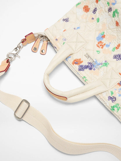 The MZ Wallace Micro Sutton in Eden Sequin Oxford is a cream quilted foldable crossbody bag with colorful floral embroidery, two handles, a detachable cream strap with silver hardware, shown on a white background.