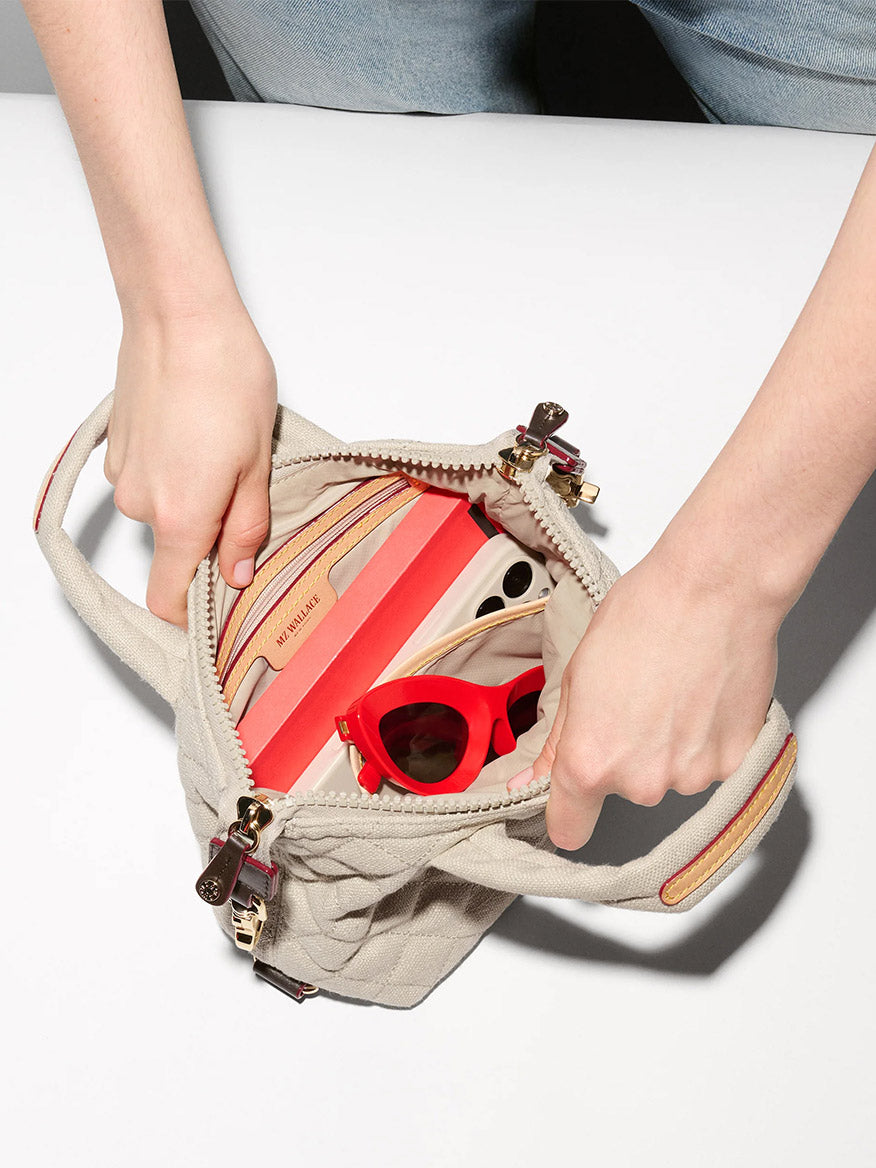 Someone holds open the MZ Wallace Micro Sutton in Sandy Linen Fabric, displaying a red wallet, red sunglasses, and a smartphone on a white surface.