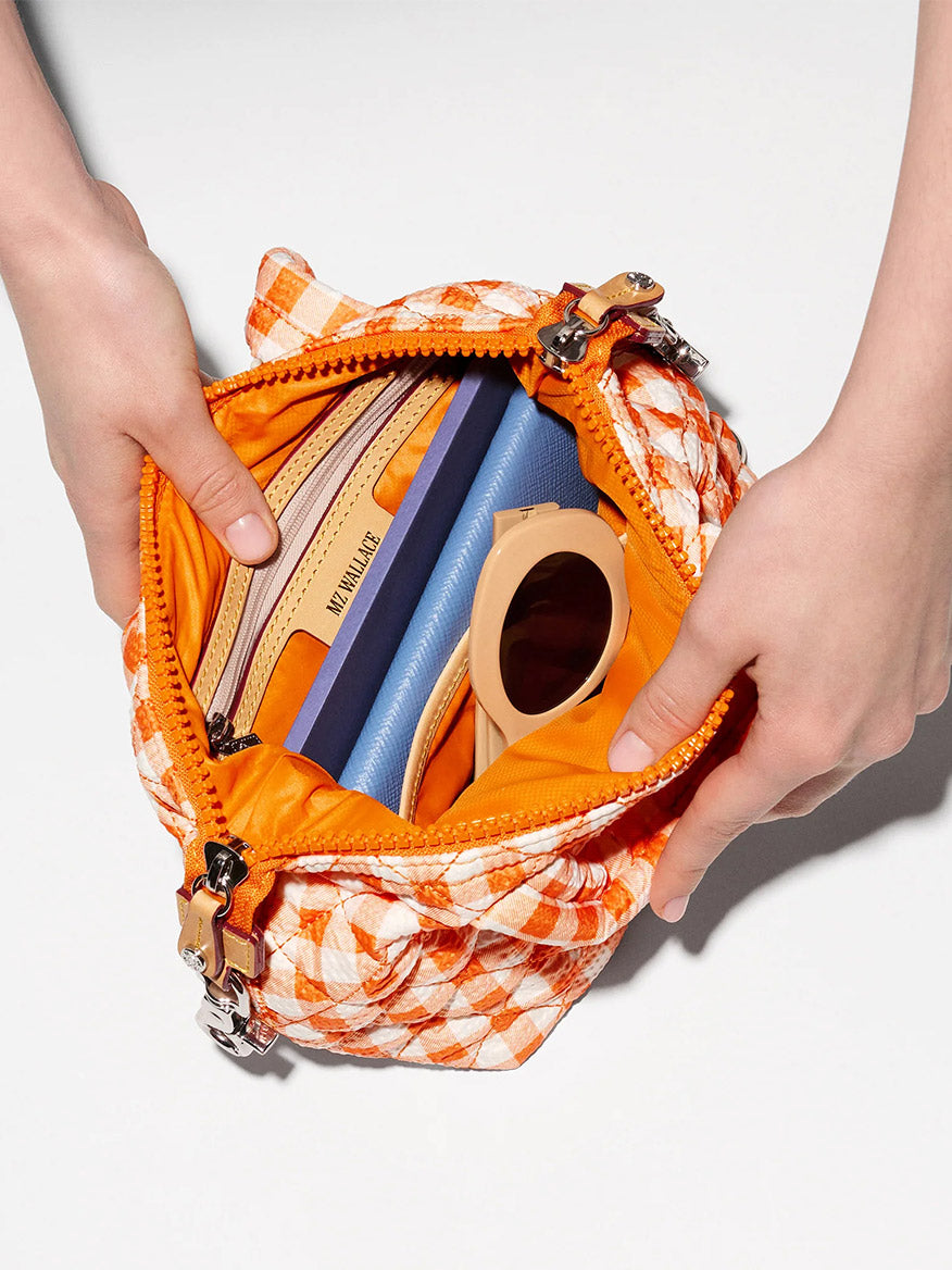 A person opens an MZ Wallace Micro Sutton in Sunset Orange Gingham Oxford, with an adjustable crossbody strap, revealing a wallet, blue notebook, beige sunglasses, and other small items inside.