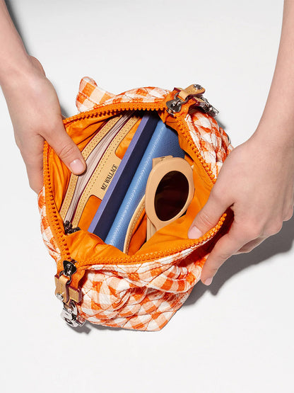 A person opens an MZ Wallace Micro Sutton in Sunset Orange Gingham Oxford, with an adjustable crossbody strap, revealing a wallet, blue notebook, beige sunglasses, and other small items inside.