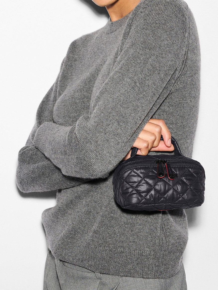 Person wearing a gray sweater and pants holds the MZ Wallace Mini Accordion Jewelry Case in Black Oxford, a small black quilted case with a handle and interior pockets, against a white background.