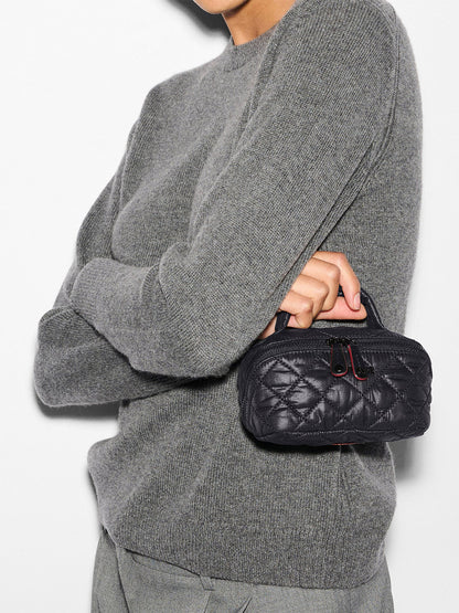 Person wearing a gray sweater and pants holds the MZ Wallace Mini Accordion Jewelry Case in Black Oxford, a small black quilted case with a handle and interior pockets, against a white background.