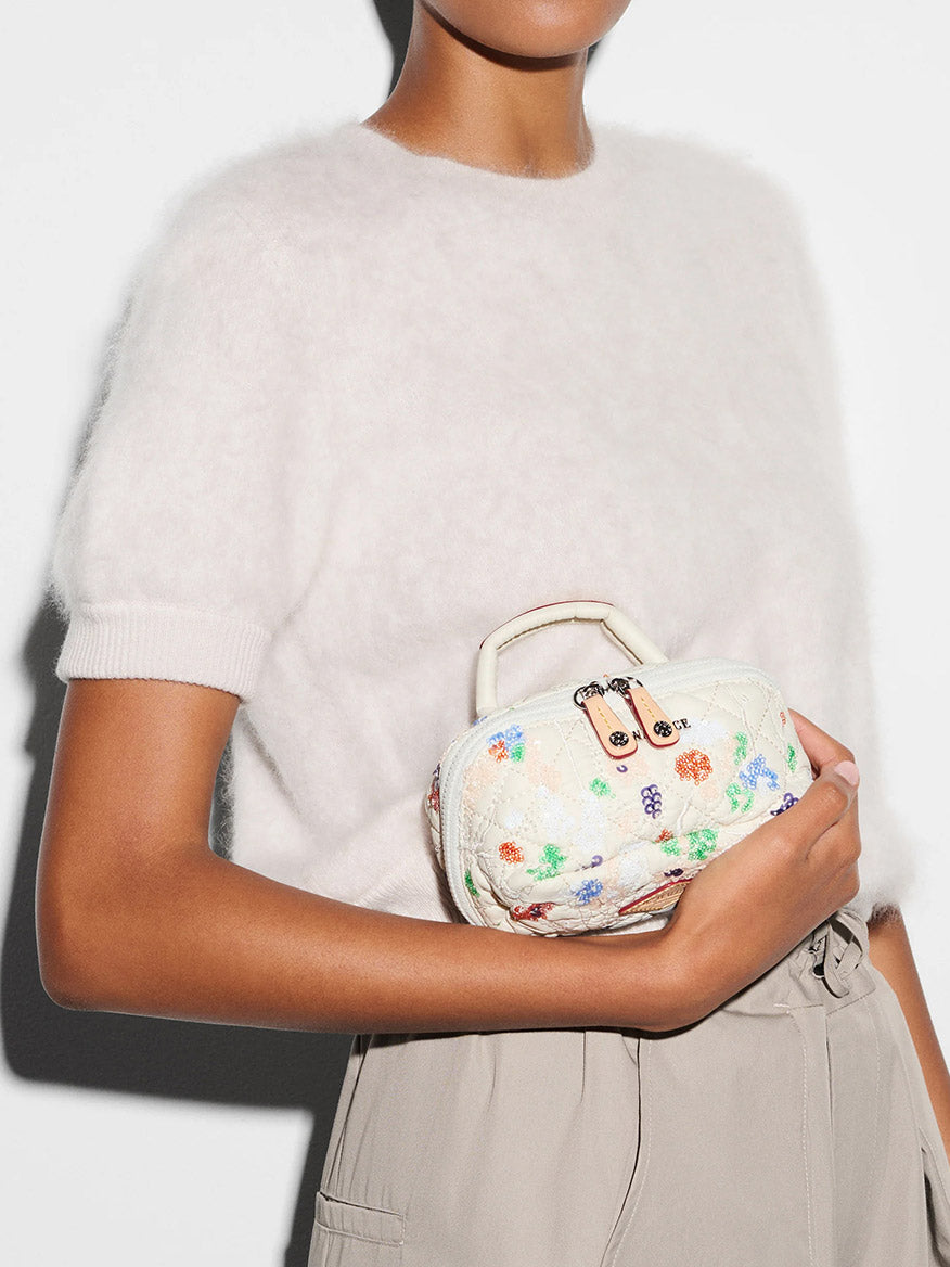A person wearing a short-sleeved, fuzzy white sweater and beige high-waisted pants holds the MZ Wallace Mini Accordion Jewelry Case in Eden Sequin Oxford.