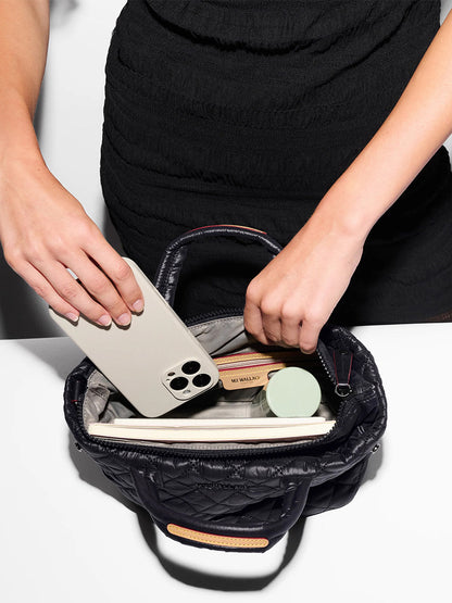 A person in a black dress puts a smartphone into the MZ Wallace Mini City Tote in Black Oxford, a small quilted bag with a crossbody strap, already holding various personal items.