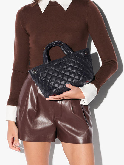 A woman in a brown long-sleeve top and brown leather shorts holds an MZ Wallace Mini City Tote in Black Oxford with a crossbody strap against a plain white background.