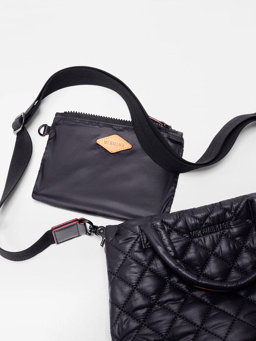 A black crossbody bag with a zippered pouch and adjustable strap lies next to an MZ Wallace Mini City Tote in Black Oxford on a white surface.