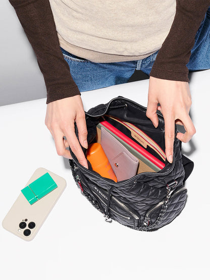A person stands over a table, opening an MZ Wallace Mini Crosby Backpack in Black Oxford filled with items, with a phone and a small green notebook nearby on the white surface.