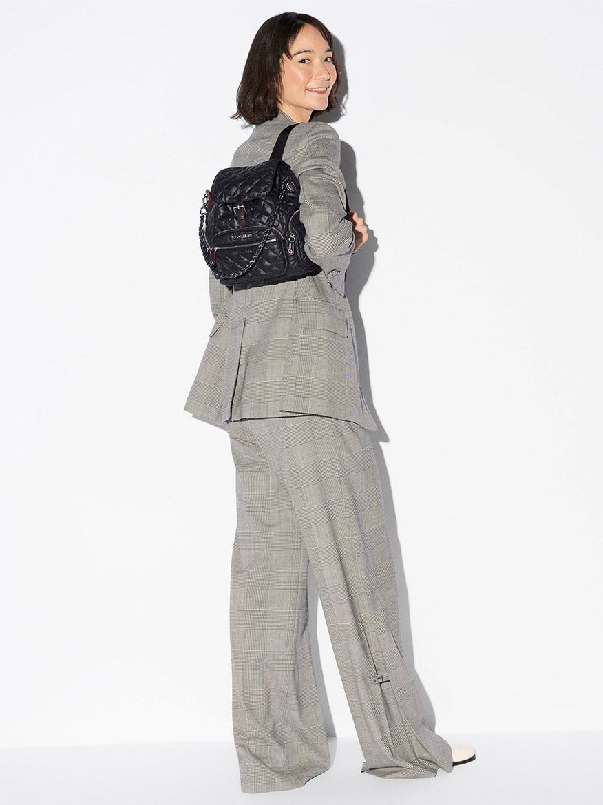 A woman in a gray plaid suit smiles, facing backward with the MZ Wallace Mini Crosby Backpack in Black Oxford slung over one shoulder against a plain white background.