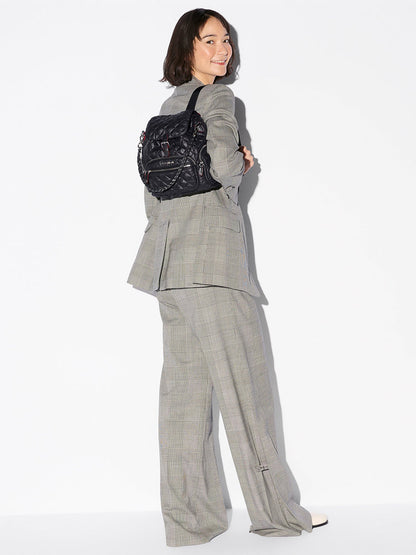 A woman in a gray plaid suit smiles, facing backward with the MZ Wallace Mini Crosby Backpack in Black Oxford slung over one shoulder against a plain white background.