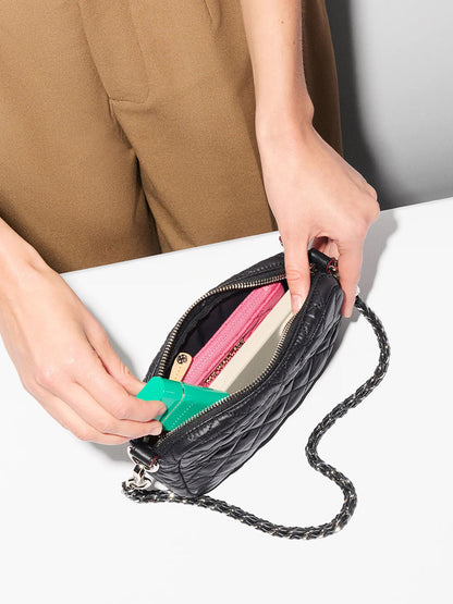 Someone puts a green item into an MZ Wallace Mini Crosby Crossbody II in Black Oxford, featuring Italian leather trim and a chain strap, along with a pink wallet and other personal belongings.