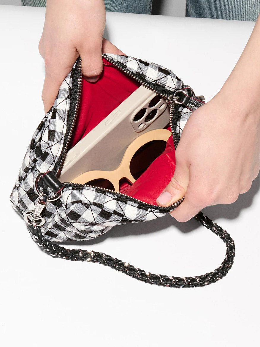 A person opens an MZ Wallace Mini Crosby Crossbody II in Black/White Gingham Oxford, revealing a smartphone and sunglasses inside.