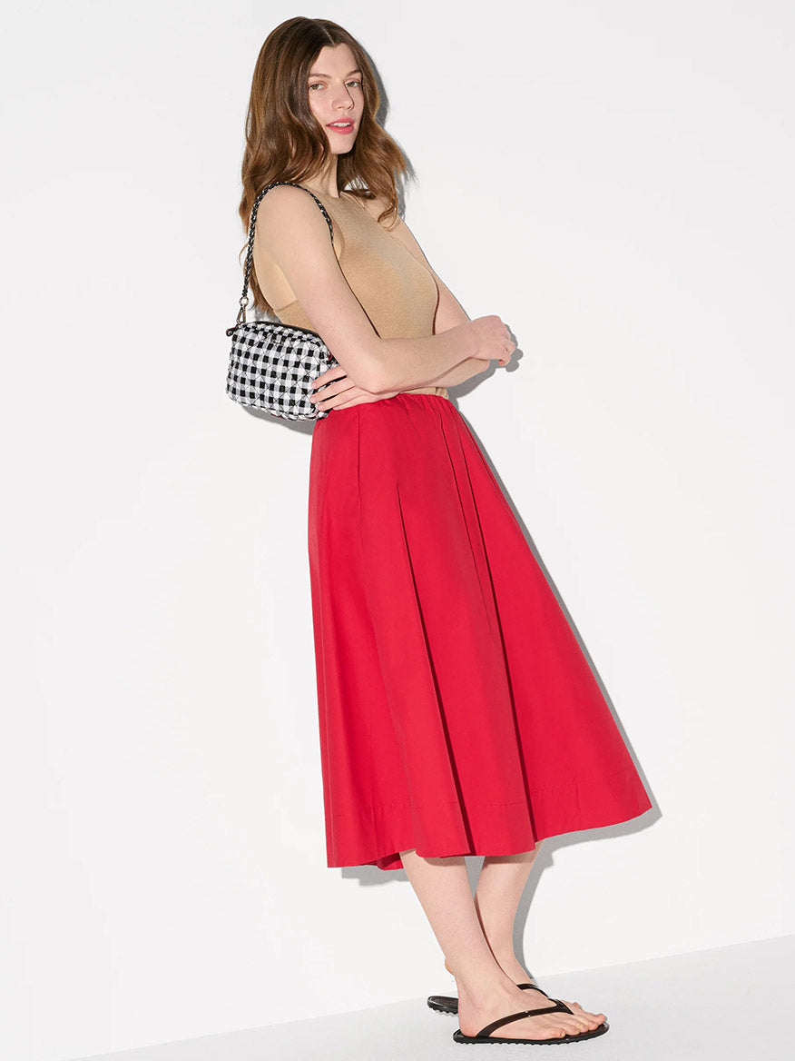 A woman stands against a plain background wearing a beige sleeveless top, red midi skirt, black flip-flops, and carrying the MZ Wallace Mini Crosby Crossbody II in Black/White Gingham Oxford with an adjustable crossbody strap.