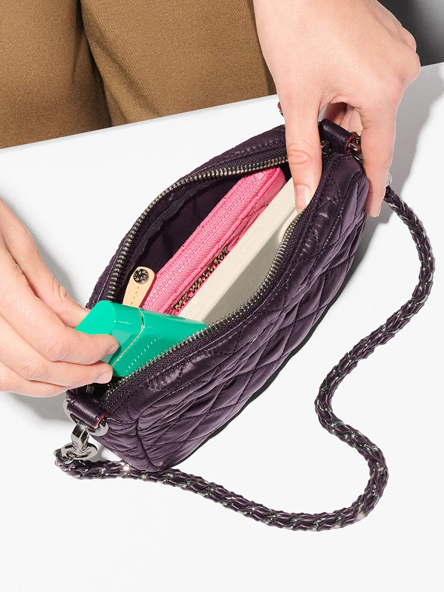A person places a green item into a small, open MZ Wallace Mini Crosby Crossbody II in Raisin Oxford, its quilted purple exterior revealing a pink wallet and a beige object on a white surface.