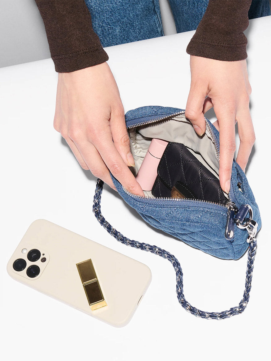 A person opens a small blue quilted pouch—an MZ Wallace Mini Crosby Crossbody II in True Blue Denim—on a white surface, with a smartphone and detachable chain strap beside it and items partially visible inside.