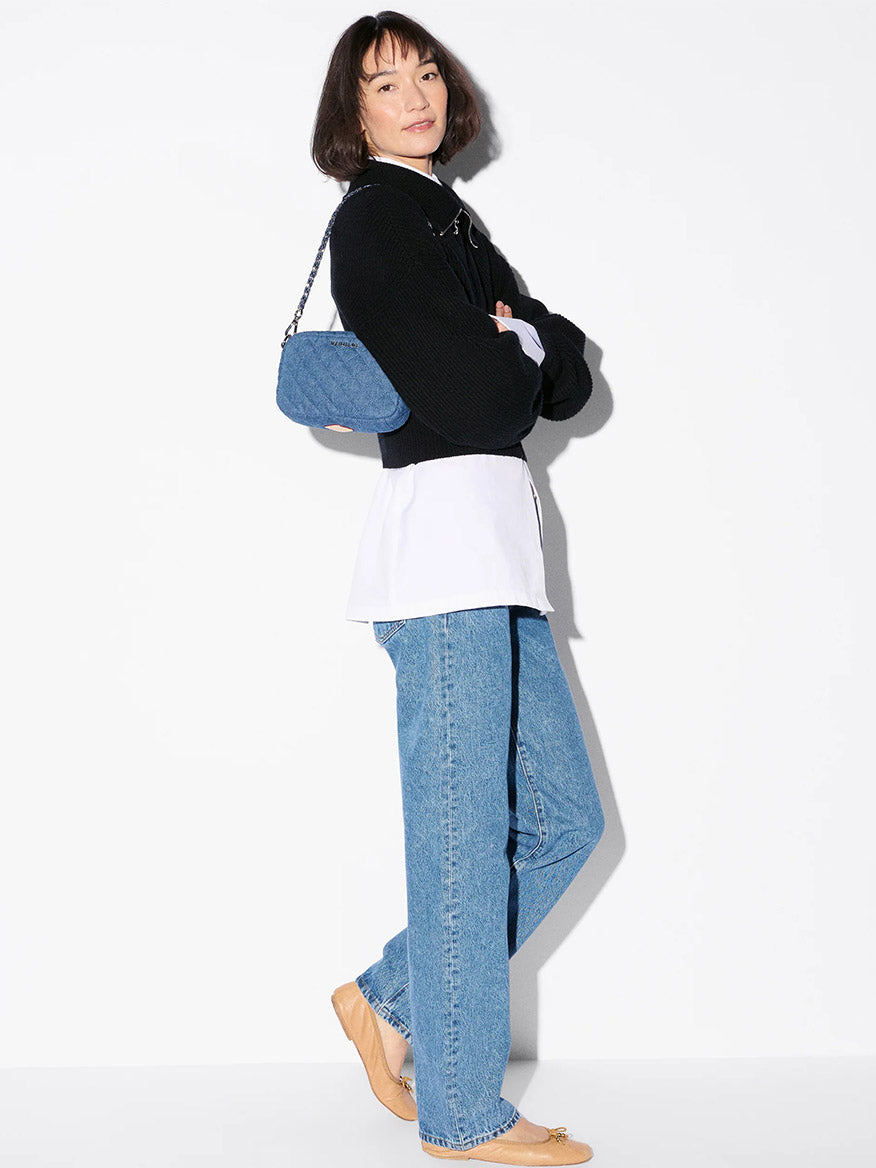 Woman stands sideways against a white background, wearing a black jacket, white shirt, blue jeans, tan flats, and carrying the MZ Wallace Mini Crosby Crossbody II in True Blue Denim with a detachable chain strap.