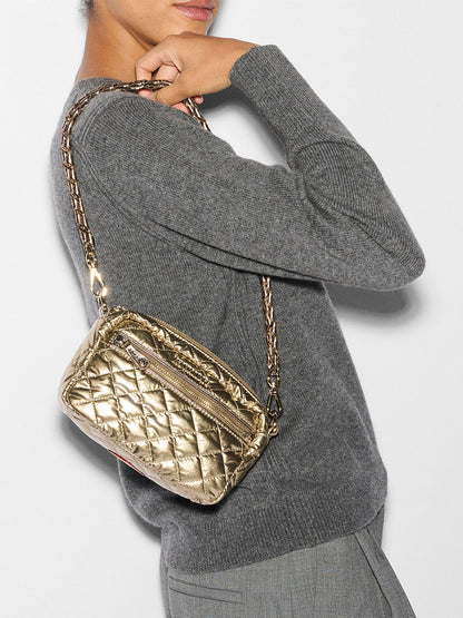 A person in a gray sweater holds a quilted MZ Wallace Mini Crosby in Antique Gold Metallic Oxford, featuring a stylish chain crossbody strap.