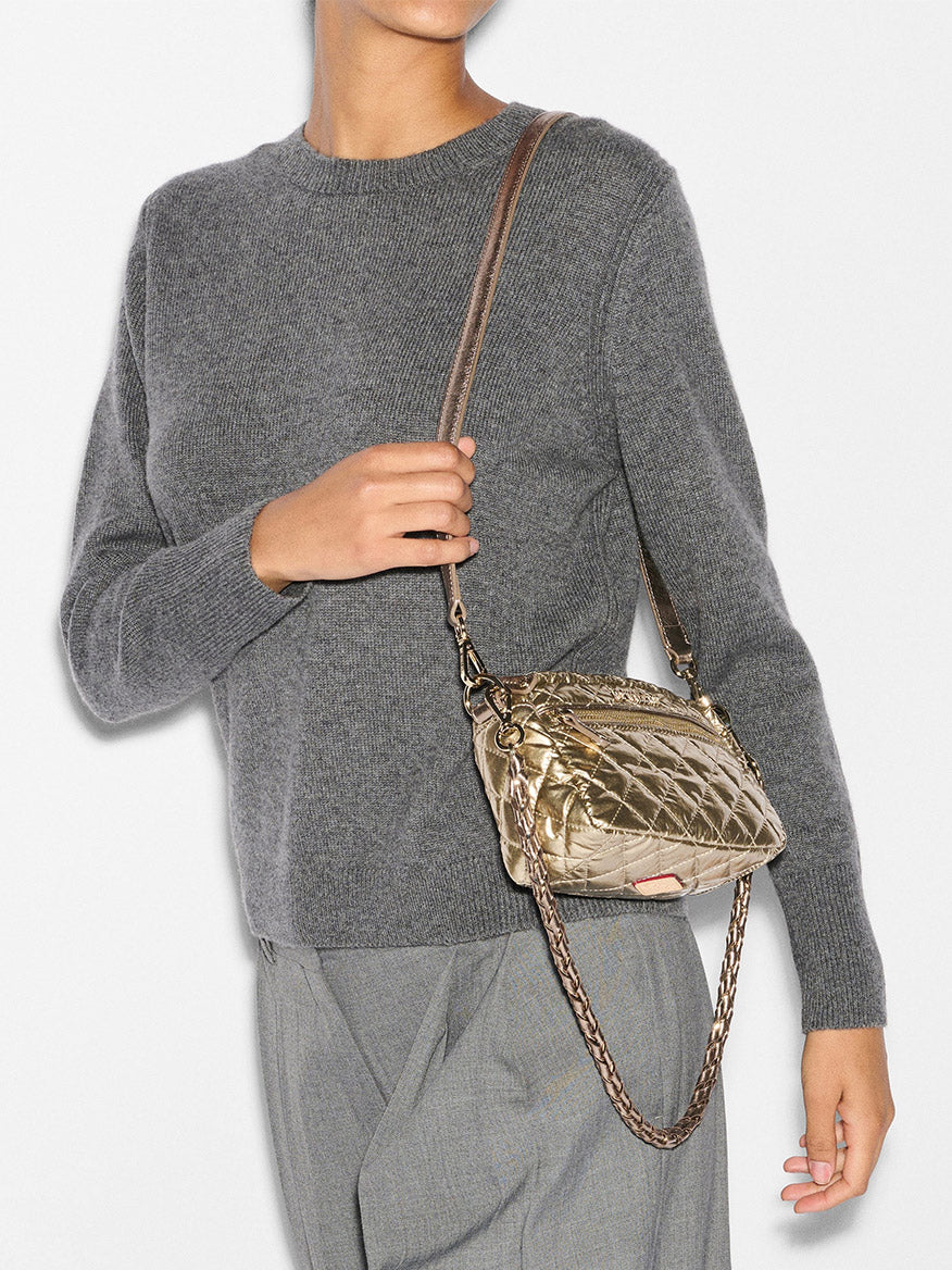 A person in a gray sweater and pants holds an MZ Wallace Mini Crosby in Antique Gold Metallic Oxford handbag with a chain strap.