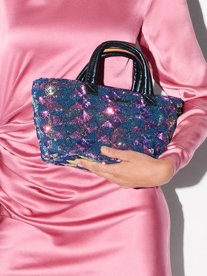 A person in a shiny pink dress holds the MZ Wallace Mini Metro City Tote in Aurora Sequin, featuring glittery blue sequins and black handles, against a plain background.