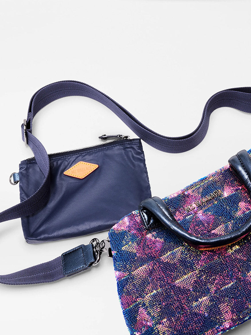 The MZ Wallace Mini Metro City Tote in Aurora Sequin, featuring a purple and pink pattern with dark handles, is displayed alongside a navy blue crossbody tote pouch with a zipper on a white surface.